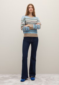 Maglione lavorato a maglia con motivi geometrici blu, marroni e crema; abbinato a jeans svasati in denim scuro e scarpe blu; tessuto testurizzato.