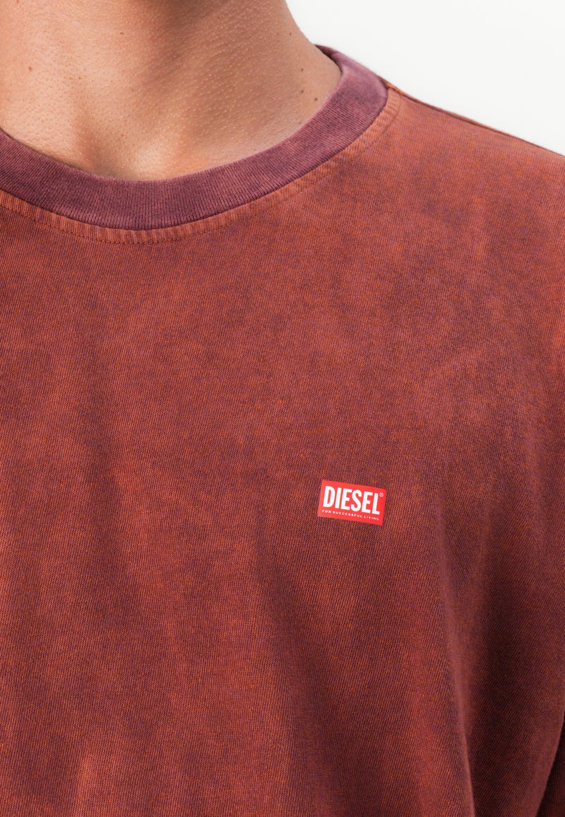 Zbliżenie osoby noszącej wyblakłą, czerwono-brązową koszulkę Diesel z małym czerwono-białym logo Diesel na klatce piersiowej.