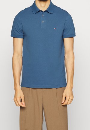 Homme portant un polo bleu à manches courtes avec des boutons et un pantalon beige, debout devant un fond clair uni.