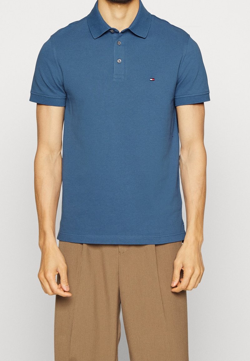 Homme portant un polo bleu à manches courtes avec des boutons et un pantalon beige, debout devant un fond clair uni.