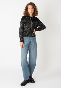 Svart stickad cardigan med intrikata vita mönster, dragkedjestängning, kragdesign, kombinerad med ljusblå raka jeanstights och svarta plattformsskor.