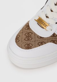 Läheltä kuvattu valkoinen tenavakenkä, jossa on ruskea monogramkankainen paneeli ja kultainen "GUESS"-nyöritagi valkoisissa nyöreissä.