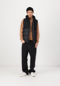 Svart puffervest med hette over en brun genser, kombinert med løse svarte jeans og hvite sneakers. Enkel design uten synlige mønstre.