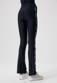 Zwarte flared leggings met een hoge tailleband. Het "BIZUU" logo staat verticaal in het wit langs de zijkant gedrukt. Glad, rekbaar materiaal. Witte sneakers.