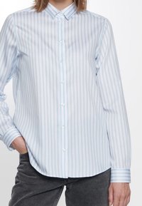 Ljusblå och vit randig skjorta med långt ärm, klassisk krage och knappslutning fram. Har en slät yta.