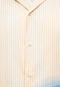 Πουκάμισο με κοντά μανίκια και κουμπιά, σε μπεζ και λευκές ρίγες. Διαθέτει γιακά και λευκά κουμπιά, με μπλε διαβαθμισμένη λεπτομέρεια στο κάτω μέρος.