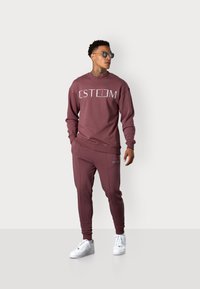 Maroones Sweatshirt und Jogginghose mit weißem "ESTEEM"-Logo, mit gerippten Bündchen und Taillenbund. Kombiniert mit weißen Sneakers und Sonnenbrille.