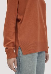 Rostorange gestrickter Pullover mit gerippten Bündchen und Saum, ausgestattet mit Seitenschlitzen. Kombiniert mit hellblauen Jeans, die eine lockere Passform zeigen.