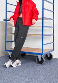 Röd puffjacka med logotyp, svarta sweatpants med logotyp och silverfärgade sportskor visar en kontrasterande färgpalett mot en blå kärra.