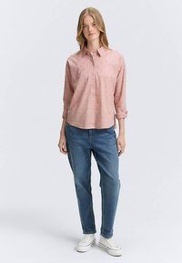 Svetloružová košeľa na zapínanie s dlhými rukávmi a malými textúrovanými bodkami; kombinovaná s modrými džínsami a bielymi teniskami.