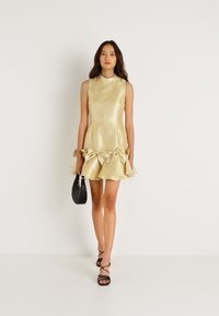 Χρυσό σατέν μίνι φόρεμα με ψηλή λαιμόκοψη, ρυτιδωτό τελείωμα και υπερμεγέθη φιόγκους στα πλάγια, συνδυασμένο με μαύρες σανδάλια με λουράκια και μια μικρή τσάντα.