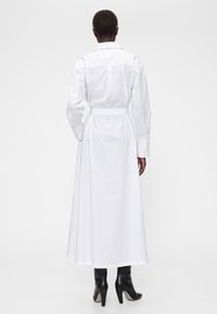 Rochie albă cu mânecă lungă, talie cu curea, guler și fustă vaporoasă. Prezintă o textură netedă din bumbac și un design minimalist.