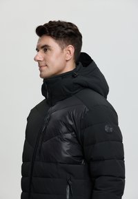 Chaqueta acolchada negra con textura de acolchado, un cuello alto, bolsillos con cremallera y capucha. Los materiales de acento incluyen paneles negros brillantes.