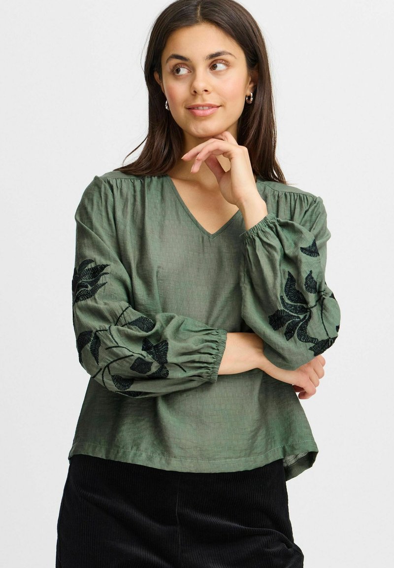 Blusa verde strutturata con scollatura a V, maniche arricciate e ricami floreali neri sulle maniche. Adatta per varie occasioni.