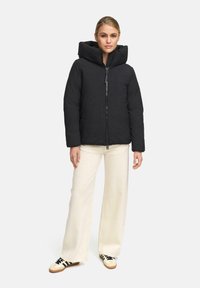 Vrouw in een zwarte gewatteerde jas met capuchon, crèmekleurige wijde broek en witte sneakers met zwarte strepen, rechtopstaand met het gezicht naar voren.