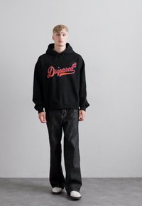 Tânăr cu păr blond, îmbrăcat într-un hanorac negru "Dsquared²", blugi largi închisați la culoare și adidași albi, stând împotriva unui zid simplu de culoare gri deschis.
