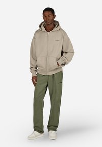 Kapučiūrinis smėlio spalvos džemperis su užtrauktuku, priekiniu kišeniu ir tonine siūle, derinamas su alyvuogių žaliųjų kelnių formos sportiniais šortais su logotipu. Baltos sportbačiai.