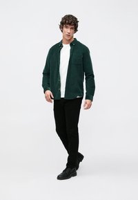 Camicia di velluto a coste verde con bottoni e taschino sul petto, indossata sopra una t-shirt bianca, abbinata a pantaloni neri e scarpe nere. Attore su uno sfondo neutro.