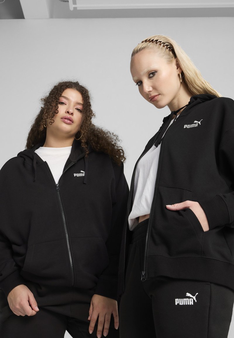 Des sweats à capuche zippés noirs avec des poches kangourou, ornés d'accents de logo Puma blancs. Un modèle porte un haut blanc en dessous, le tout associé à des joggers noirs.
