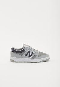 Šedé a černé tenisky New Balance se šňůrkami, perforovanou špičkou a bílou podrážkou na jednobarevném bílém pozadí.