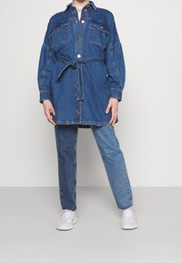 Jeansskjolklänning med långa ärmar, knappstängning, två bröstfickor och knytbälte i midjan. Bärs med ljusare jeansbyxor och vita sneakers.