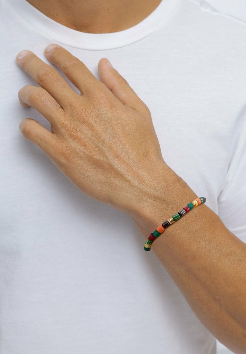 Männliche Hand ruht auf der Brust und trägt ein buntes Perlenarmband mit grünen, roten, orangefarbenen und goldenen Perlen vor einem weißen Hemd.