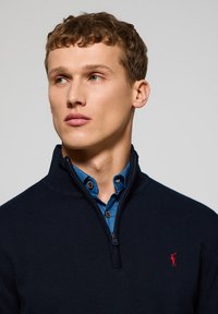 Suéter azul marino con cremallera y cuello alto, superpuesto a una camisa azul con botones visibles, que presenta un pequeño logo rojo en el pecho.