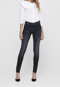 Camisa blanca de botones con un gran collar arrugado, combinada con jeans negros ajustados y tacones negros; tejido suave con un ligero brillo.