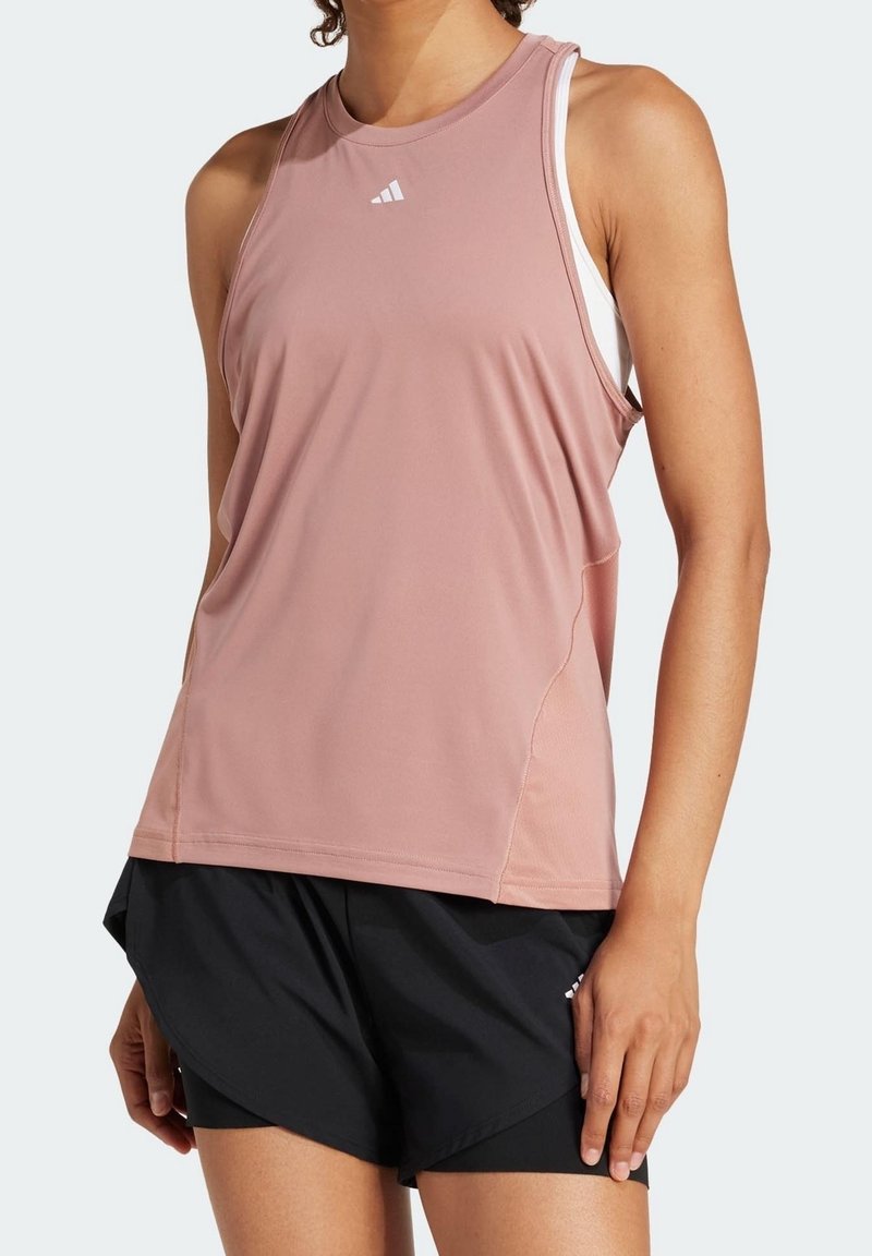Femme portant un débardeur de sport rose clair sans manches et un short de course noir, debout devant un fond uni.