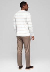 Muž v bílých dlouhých rukávech košili s tenkými béžovými pruhy a šedých kalhotách, stojící a otočený zády k jednoduchému šedému pozadí.