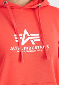 Sudadera roja con capucha ajustable, con un logo blanco y el texto "ALPHA INDUSTRIES" en la parte frontal, textura de tela suave.