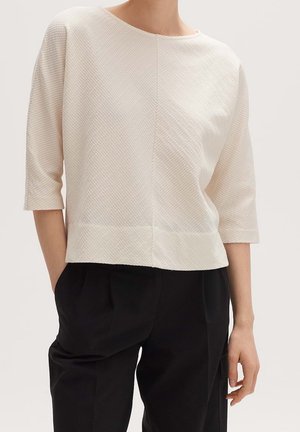 Vrouw draagt een crèmekleurige blouse met textuur, driekwart mouwen en een ronde hals, gecombineerd met zwarte, hooggetailleerde broek met zakken.