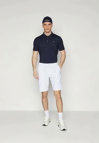 Jūras spēku polo krekls ar logo, baltas šorti un baltas sporta apavi. Modelis valkā tumši zilu beisbola cepuri. Vienkāršs, sportisks dizains ar pieguļošu formu.