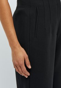 Pantaloni neri su misura con una texture liscia. Presentano tasche laterali e una parte anteriore piatta, evidenziata da dettagli di cucitura discreti.
