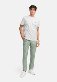 Grå bomullströja med en grön cirkulär logotyp, kombinerad med ljusgröna linbyxor och vita sneakers, visad på en modell.