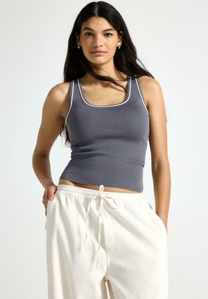 Ung kvinde med langt mørkt hår iført en grå, ærmeløs ribbet tanktop og hvide bukser med snor, stående med hænderne i lommerne.