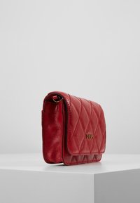 Punainen nahkalaukku, jossa on tikattu kuosi, kultainen logo ja läppäkiinnitys. Kompakti, suorakulmainen muotoilu, jossa on sileä pinta.