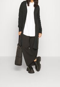 Dlouhý černý kardigan přes bílou košili, v kombinaci se širokými černými kalhotami a robustními černými botami, držící tmavou prošívanou tašku.