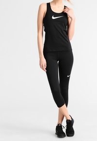 Débardeur de sport noir avec un logo Nike blanc, associé à des leggings capri noirs et des baskets noires. Tissu lisse, coupe ajustée.