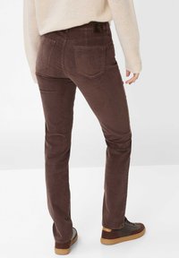 Pantaloni in velluto a coste marroni con un design sagomato, dotati di tasche posteriori e tessuto testurizzato, abbinati a sneaker marroni scuri.