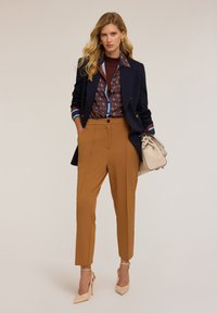 Blazer navy sopra una camicia a fantasia con accenti marroni e blu, pantaloni beige e tacchi alti beige, abbinati a una borsa chiara.