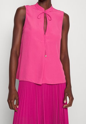 Mouwloze felroze blouse met een knoopsluiting bij de hals en een hoge taille geplooide magentakleurige rok, gedragen door een persoon met een donkere huid.