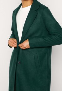 Grüner Wollmantel mit Reverskragen, Einzelknopfverschluss und Seitentaschen, getragen über einem weißen, strukturierten Pullover. Glatte Stoffoberfläche.