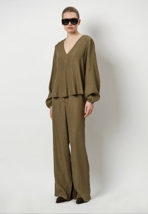 Olijfgroen textuuroutfit met een V-hals, lange ballonmouwen en wijdlopende broeken met een lichte glans. Donkere zonnebril maakt de look compleet.