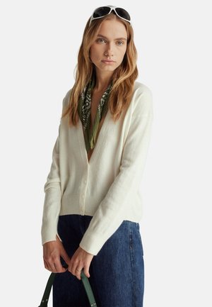 Cardigan avorio con polsini e orlo a coste, dotato di bottoni sul davanti. Abbinato a jeans scuri e a una sciarpa verde con motivo.