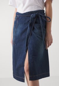 Denimová zavinovací sukně v tmavě modré barvě s uvázaným pasem, která má přední rozparek, viditelné švy a hladkou strukturu.