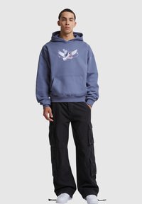 Hooded sweatshirt in light blue with a graphic of a bird design on the chest, paired with black cargo pants and white sneakers.