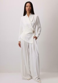 Bela zavita bluza z dolgimi rokavi, z nagubanim dizajnom in zlato poudarkom v predelu pasu, kombinirana z belimi nagubanimi širokimi hlačami.