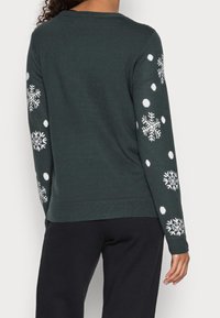 Pull en vert foncé avec des motifs de flocons de neige et de points blancs sur les manches, col et poignets côtelés, fabriqué en matériau tricot doux.