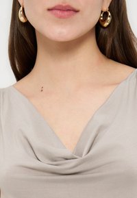 Γυναίκα που φοράει χρυσούς κρίκους στα αυτιά και μπλούζα μπεζ με draped λαιμόκοψη, με δύο μικρούς σπίλους ορατούς στον λαιμό.